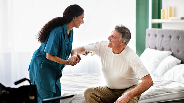 A healthcare worker with curly hair helps an older man in a white shirt while he sits on the edge of a bed. They are engaged and smiling in a well-lit bedroom setting.