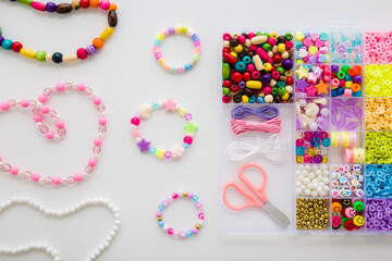 Bracelet, necklaces, scissors, elastic strings and case with colorful different plastic parts on light gray table background. Closeup. Creating beaded jewellery for little child girl. Top down view.