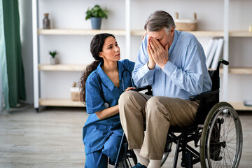 Naklejka premium A woman with dark hair wearing a blue uniform sits beside a man with gray hair in a wheelchair. The man covers his face, indicating strong emotions.