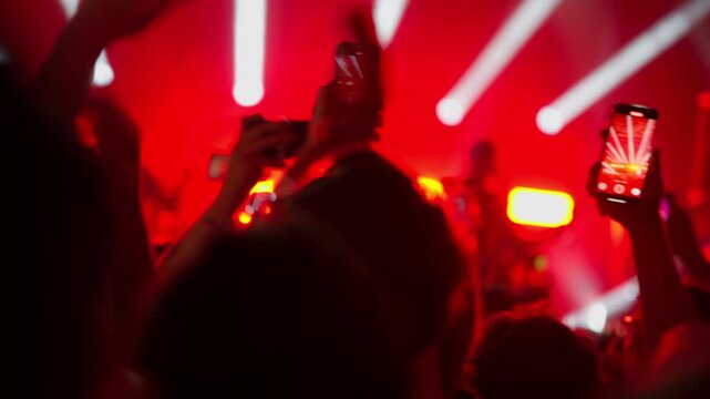Silhouetted crowd at a music festival or concert. People hold up smartphones, recording the show under dramatic red stage lights. Energetic, modern nightlife and live event atmosphere footage.