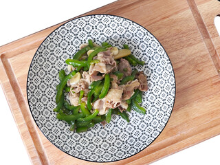 Top-down still of a plate of stir-fried green bell pepper and thinly sliced pork, served in a patterned bowl on a wooden cutting board