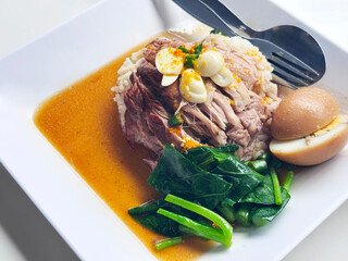 Thai-style braised pork and rice dish, tender pork sits atop jasmine rice, topped with sliced garlic and chopped chilis, accompanied by blanched greens and a halved stewed egg. Isolated image on white