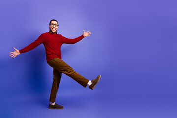 Fototapeta premium Happy young man in a red sweater balances on one leg against a violet backdrop for a lively fashion lifestyle stock photo