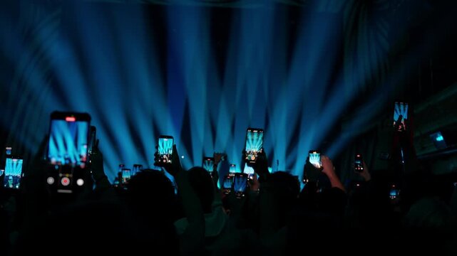 Footage of a silhouetted crowd at a concert holding up phones to record the show. The camera slowly zooms in on the hands and screens, with dramatic blue spotlights lighting the stage.