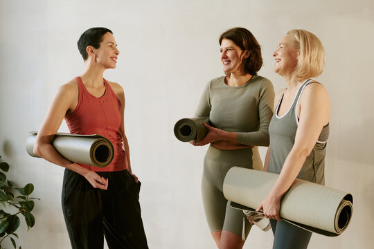 Three middle aged Caucasian women standing together holding yoga mats, smiling and talking before fitness class, showing relaxed body language and friendly interaction