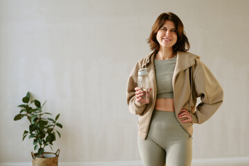 Fototapeta na wymiar Portrait of young adult Caucasian woman smiling at camera holding reusable water bottle standing indoors with hand on hip showing healthy lifestyle and confidence