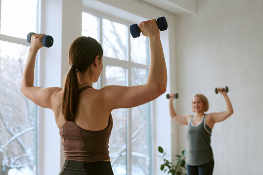 Young adult Caucasian woman and middle aged Caucasian woman exercising with dumbbells in bright room, both raising arms and performing strength training workout together near large windows