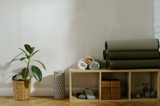 Yoga studio interior showing rolled mats, foam rollers, cork blocks, dumbbells and resistance bands arranged on wooden shelf with potted plant on floor against plain wall, no people visible