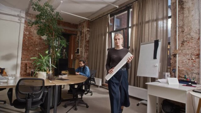 Young Caucasian female architect rolling out printed floor plan on table while working with diverse colleagues in bright, spacious office environment