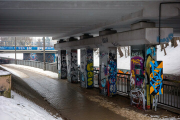Fototapeta premium Warsaw, Masovian Voivodeship, Poland - January 27, 2026: A wide-angle shot of a pedestrian walkway beneath a concrete bridge structure heavily covered in vibrant graffiti art