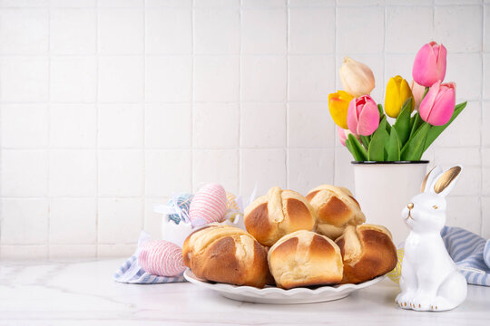 Traditional Easter hot cross buns. Traditional spring holiday Easter baking pastry &ndash; hot cross buns. On white background with sunlight, Easter eggs, bunny rabbits and tulip flower bouquet 