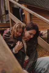 Mother and daughter hugging on the steps of a wooden staircase. Cheerful girls in the country in the fall. A little girl hugs her mother from behind. Rainy weather. Vertical photo.
