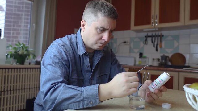 A man in the kitchen tests tap water quality with a test strip. Home testing for drinking water safety, pH, chlorine, and chemical contaminants.