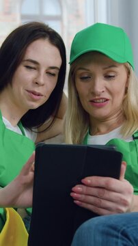 Good-looking joyful professional young women-workers of cleaning company sitting on sofa and watching something interesting on tablet device during cleaning up client's flat