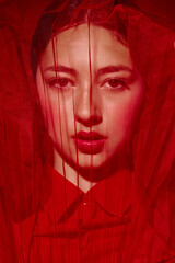 Close-up portrait of a young woman in red makeup and clothing, partially veiled with red fabric....