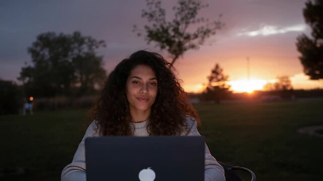 Smiling woman wearing light sweater sitting behind laptop in park at dusk, camera focusing, working