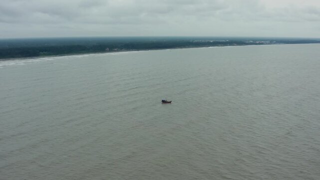 Aerial drone captures lone fishing boat adrift in grey ocean waters parallel to distant tree-lined Talsari Beach under expansive cloudy skies in Odisha, India.