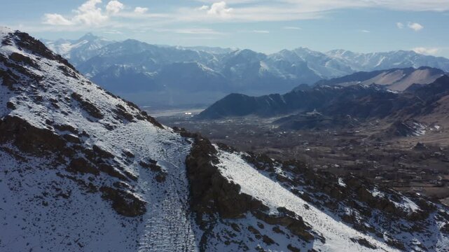 Drone reveal pans from snow-dusted ridges over Leh city valley nestled amid stark mountains with Stok Kangri range dominating the horizon under clear skies.