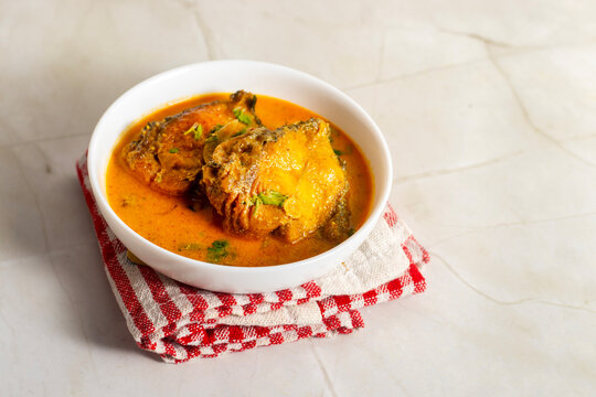 White bowl holds Indian katla/catla fish curry Bengali style with golden gravy, tender fish pieces, onion slices, and herbs, styled on marble surface with red cloth background.
