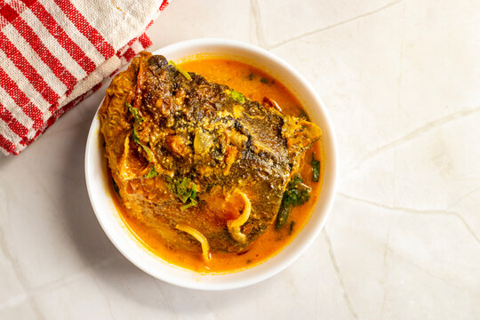 Top view of Bengali catla fish curry in white bowl, rich mustard gravy, herbs and onions, served on marble surface with red cloth.
