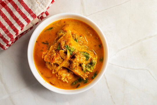 White bowl holds Indian katla/catla fish curry Bengali style with golden gravy, tender fish pieces, onion slices, and herbs, styled on marble surface with red cloth background.
