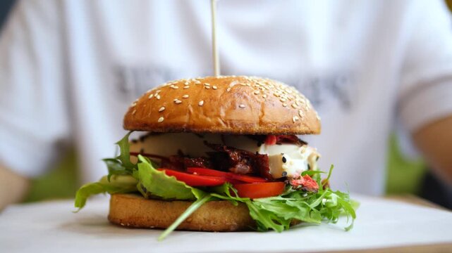 Chef presenting a freshly made gourmet burger with a sesame seed bun, juicy patty, fresh arugula, tomatoes, and cheese, ready to be served on a white plate in a restaurant setting