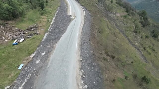High-angle drone footage shows a narrow asphalt road carving through steep, green, mountainous terrain. Evidence of disarray (tents, debris) is visible on the slopes bordering the road.