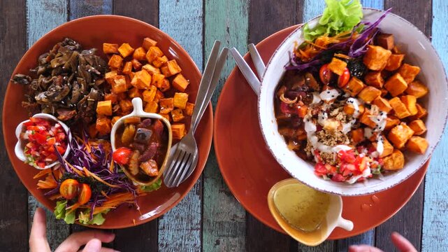 Waiter's hands placing two vibrant and appetizing vegan lunch plates on a rustic colorful wooden table, featuring sweet potatoes, pulled mushrooms, fresh salad, and delicious sauces