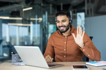 Confident bearded man wearing a headset and smiling, sitting at a desk in a modern office space,...