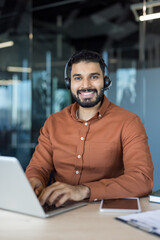 Obraz premium Smiling male call center agent wearing a headset, providing online assistance and technical support while typing on a laptop in a modern corporate office environment