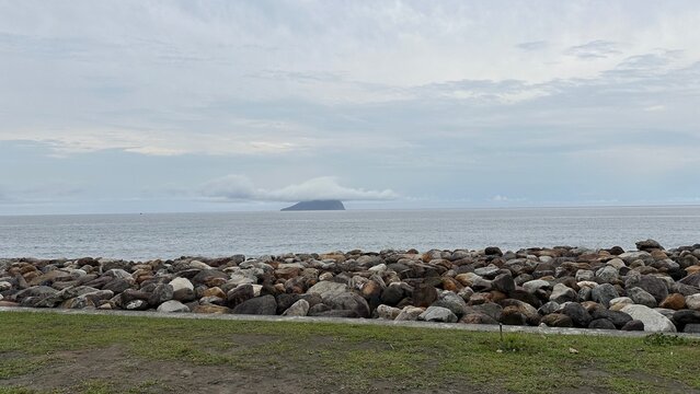 A tranquil view of Guishan Island in Taiwan, A rocky shoreline foreground with colorful stones, leading to a calm ocean and a distant island partially covered by clouds under a gray sky.