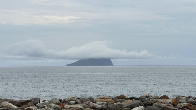 A tranquil view of Guishan Island in Taiwan, A rocky shoreline foreground with colorful stones, leading to a calm ocean and a distant island partially covered by clouds under a gray sky.