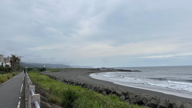 Dakeng Beach in Toucheng, Taiwan. A winding coastal path lined with grass and concrete barriers, leading to a gray sandy beach under a cloudy sky.