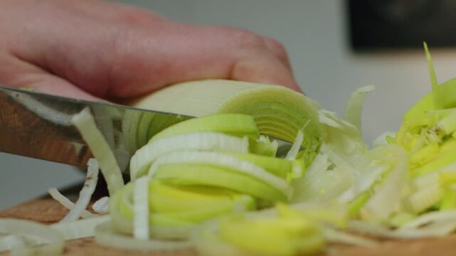 A hand cuts leeks on a wooden board in a kitchen. The green parts of the leeks are neatly arranged
