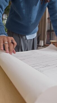 Vertical shot of Biracial male architect rolling out paper building plan on table reviewing project details in modern office workspace