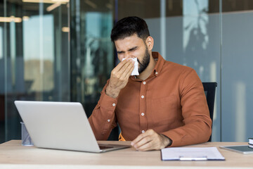 Obraz premium Man feeling unwell and sneezing into a paper tissue while working on a laptop at an office desk, illustrating illness, allergies, or a common cold affecting productivity