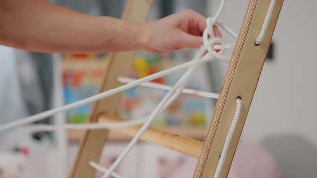 Person ties knots on a wooden ladder during an indoor activity in a bright room with colorful toys in the background