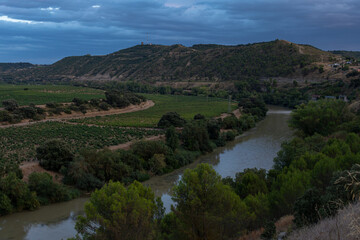 Elevated view of the Ebro river flowing past lush vineyards and rolling hills under a dramatic twilight sky in La Rioja, Spain