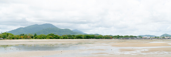 Panoramic view of a tropical beach at low tide with wet sand and puddles. Lush green trees line the shore with rolling mountains under a cloudy sky in the background. Calm coastal landscape.