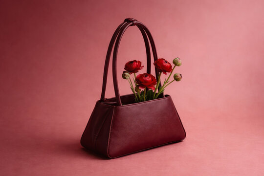 Burgundy leather handbag, sculptural handles against soft rose background. Red ranunculus flowers emerging from interior, femininity, gift mood, fashion still life. Valentine&rsquo;s Day concept.