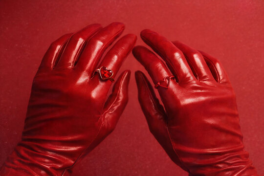 Red leather gloves with heart-shaped rings against deep red background. Symmetrical hand gesture, tactile surface, intimate mood, fashion still life with focus on touch and emotion. Valentine&rsquo;s Day