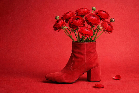 Red suede ankle boot holding bouquet of ranunculus flowers against textured red background. Fashion still life, bold monochrome palette, playful romance, editorial. Valentine&rsquo;s Day concept.