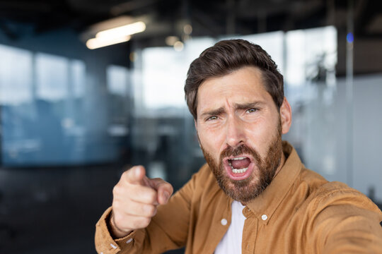 Frustrated man expressing anger and disagreement, shouting and pointing finger at a camera while standing in a modern office environment, symbolizing conflict and communication issues