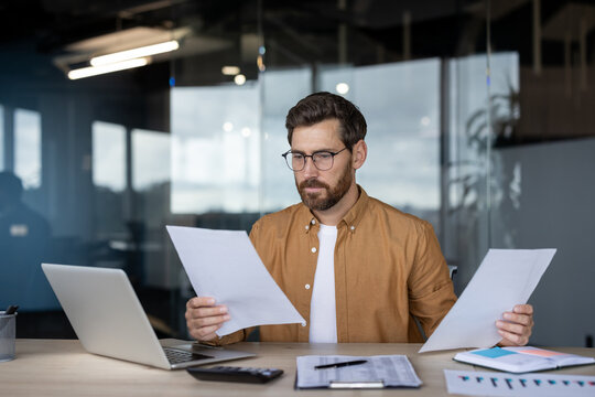 Businessman focusing intently while analyzing paperwork and financial reports at his office desk, comparing information on documents with data displayed on a laptop screen