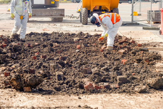 Two workers in protective gear remove asbestos from contaminated ground filled with bricks at construction site