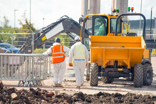 Two workers in safety suits carry out asbestos removal from contaminated ground near construction equipment
