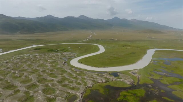 Aerial view of a winding river flowing through verdant green fields, creating a stunning contrast against the backdrop of distant mountains, Ba Yin Guo Leng Meng Gu Zi Zhi Zhou, China.