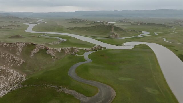 Aerial view of a river snaking through green grassland with hills under a cloudy sky, Ba Yin Guo Leng Meng Gu Zi Zhi Zhou, Xin Jiang Wei Wu Er Zi Zhi Qu, China.