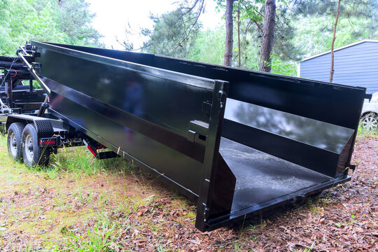 Waste dump trailer is stationed in woods close to house on clear day.