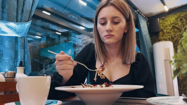 Business woman is eating an organic salad meal at a restaurant. She is dressed in black has a spoon in her hand. The table is set with a plate of food and a cup. The atmosphere is casual and relaxed